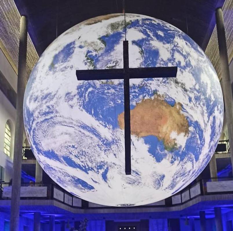 In einem Raum schwebende Weltkugel, davor ein schwebendes Kreuz