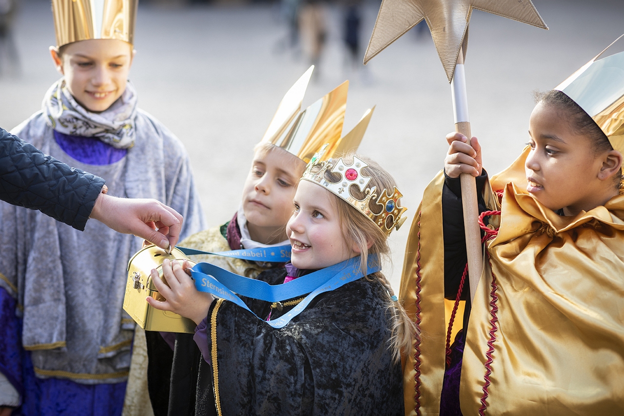 Vier lächelnde Kinder, verkleidet als Sternsinger mit goldenen Kronen und Umhängen, halten eine Sammelbüchse, in die eine Hand gerade eine Münze einwirft. Ein Kind hält einen großen Stern. (Autor / Kindermissionswerk)